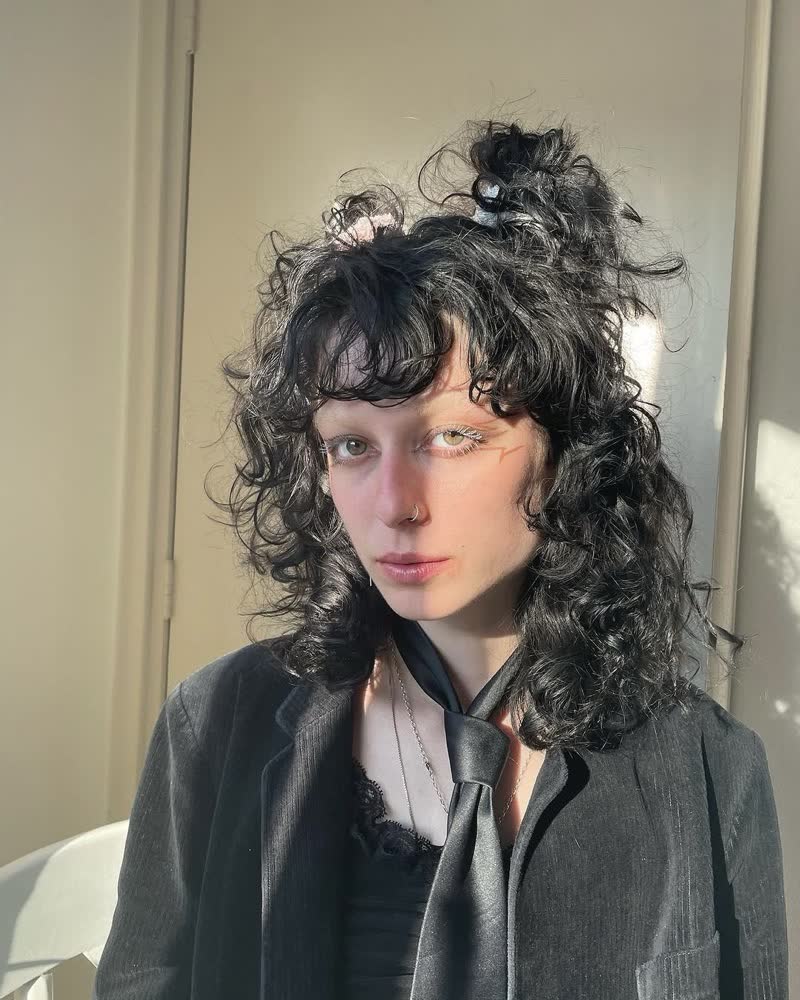 cyberbirdxx seated against a pale door in a grey blazer over black, half in sun, looking straight out