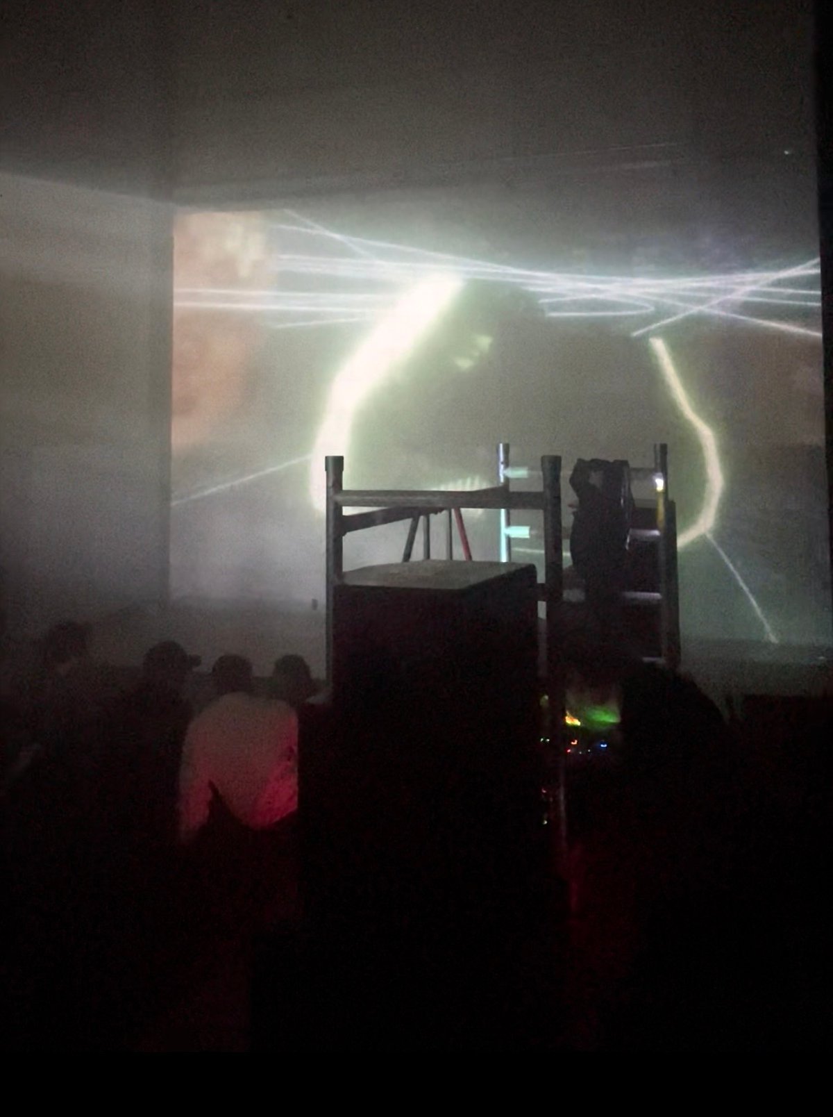 a room in haze · light traces projected across the back wall, the crowd and a scaffold rig in silhouette