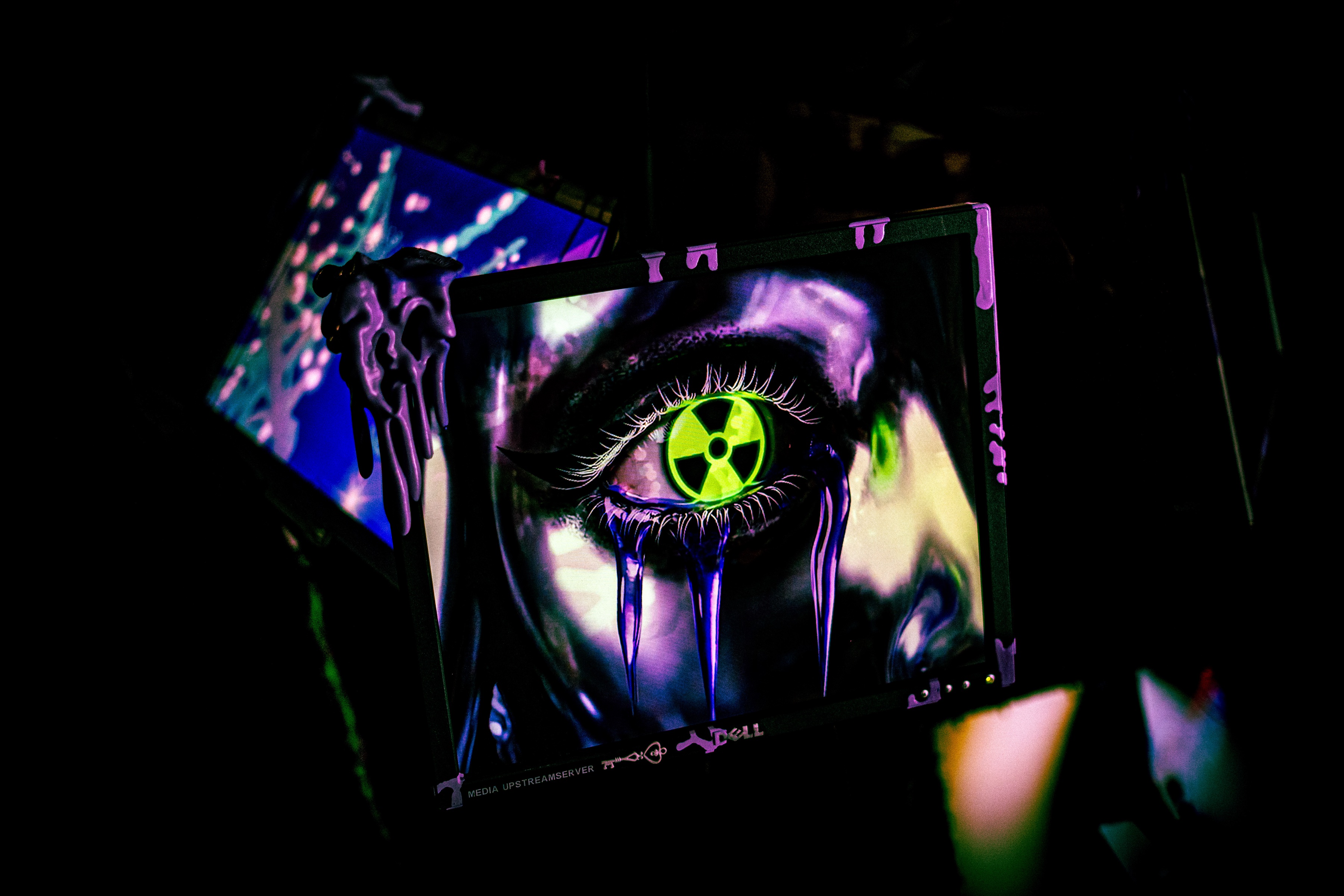 a dell monitor tilted in the dark, cyberslime drooling from its top corner, the screen showing a large painted eye with a green radiation-trefoil iris weeping violet chrome tears, another screen glowing blue behind it