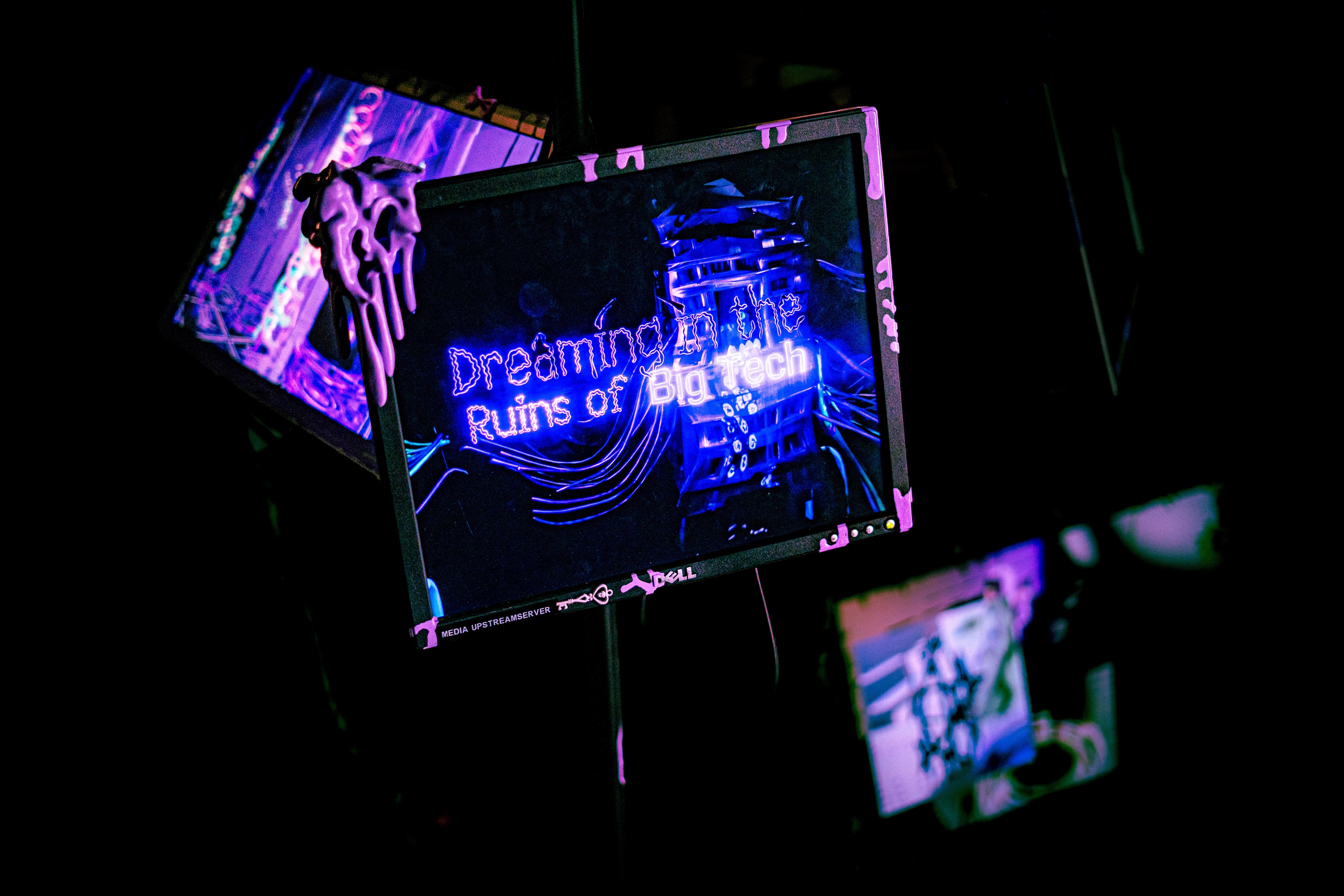 a canted dell panel with pink slime running off the frame, the words dreaming in the ruins of big tech in violet and white over a dark tower of scaffold pole, two more screens glowing blue and pink behind