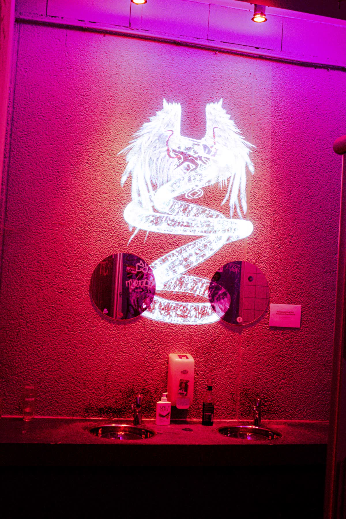 the winged figure projected on the bathroom wall above the sinks, the whole room flooded magenta