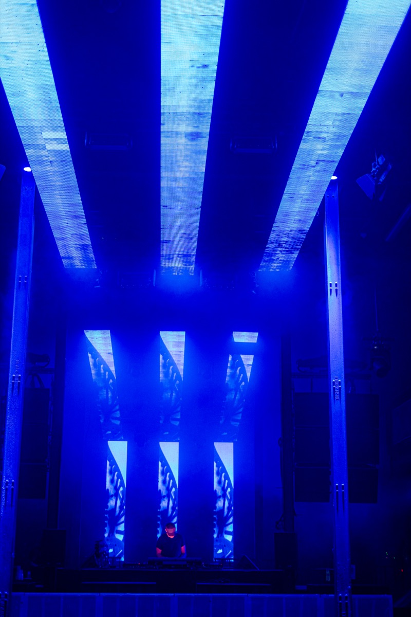 led panels lit blue, branching thread-like patterns like strands of dna running down each screen, a dj lit small below.