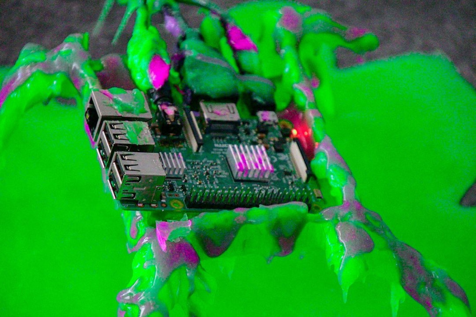 a raspberry pi board almost buried in bright green and magenta cyberslime, drips hanging off every edge, a red status light glowing through the coating.